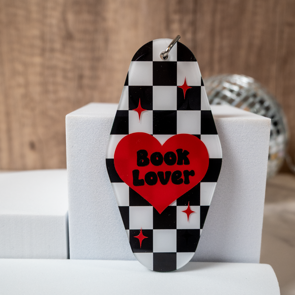 Decorative keychain with checkered pattern and red heart-shaped 'Book Lover' text on a white stand.