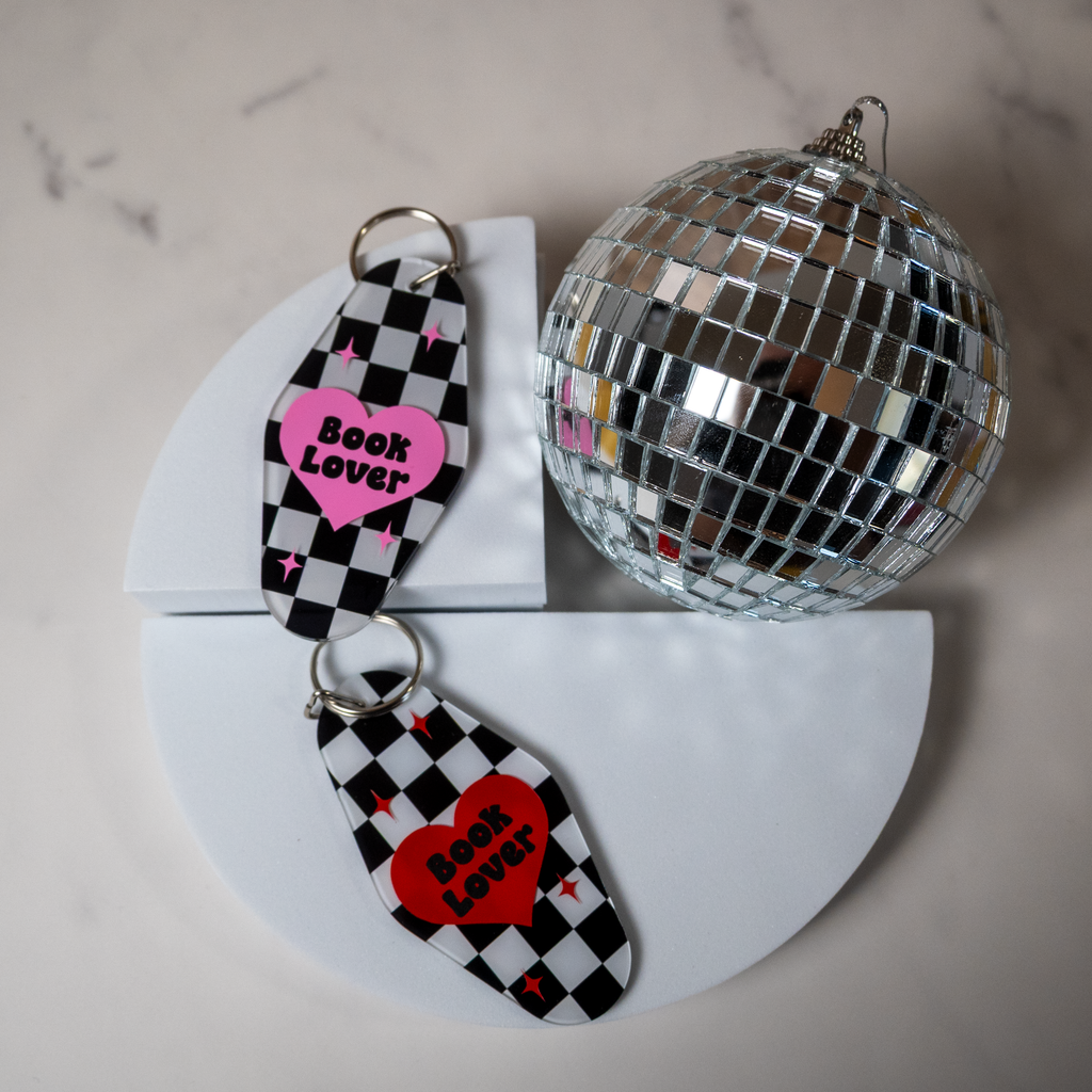 Two keychains with heart designs and 'Book Lover' text next to a disco ball on a marble surface.