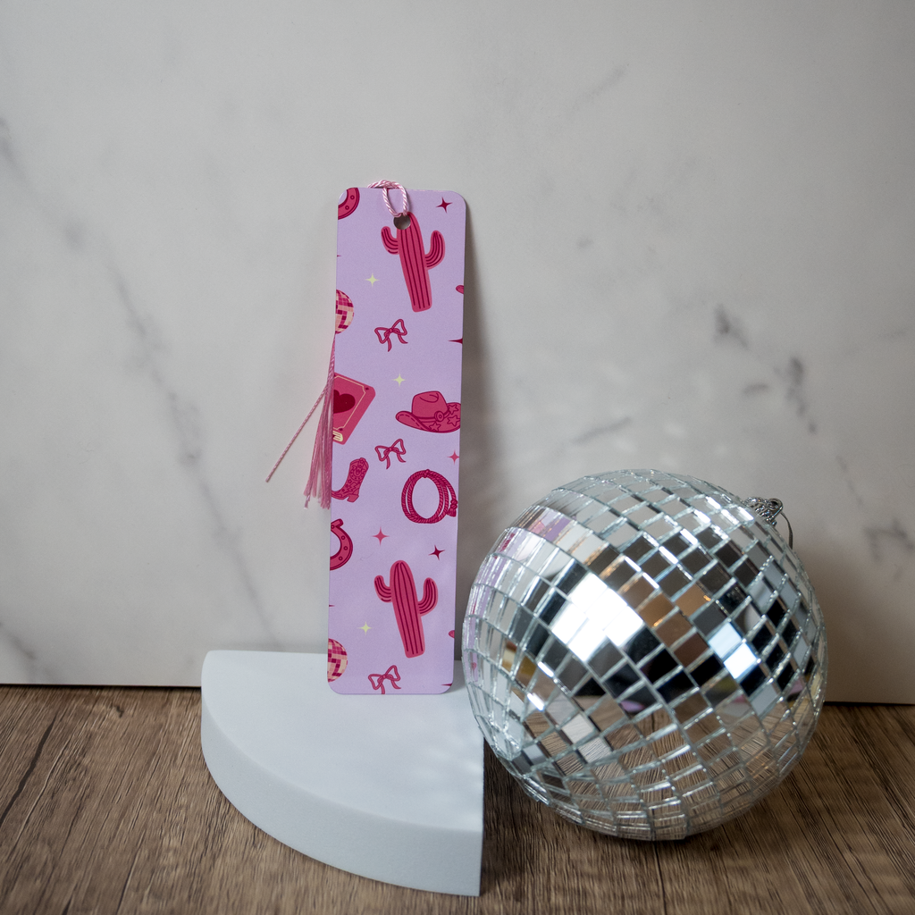 Pink bookmark with cactus pattern next to a silver disco ball on a wooden surface.