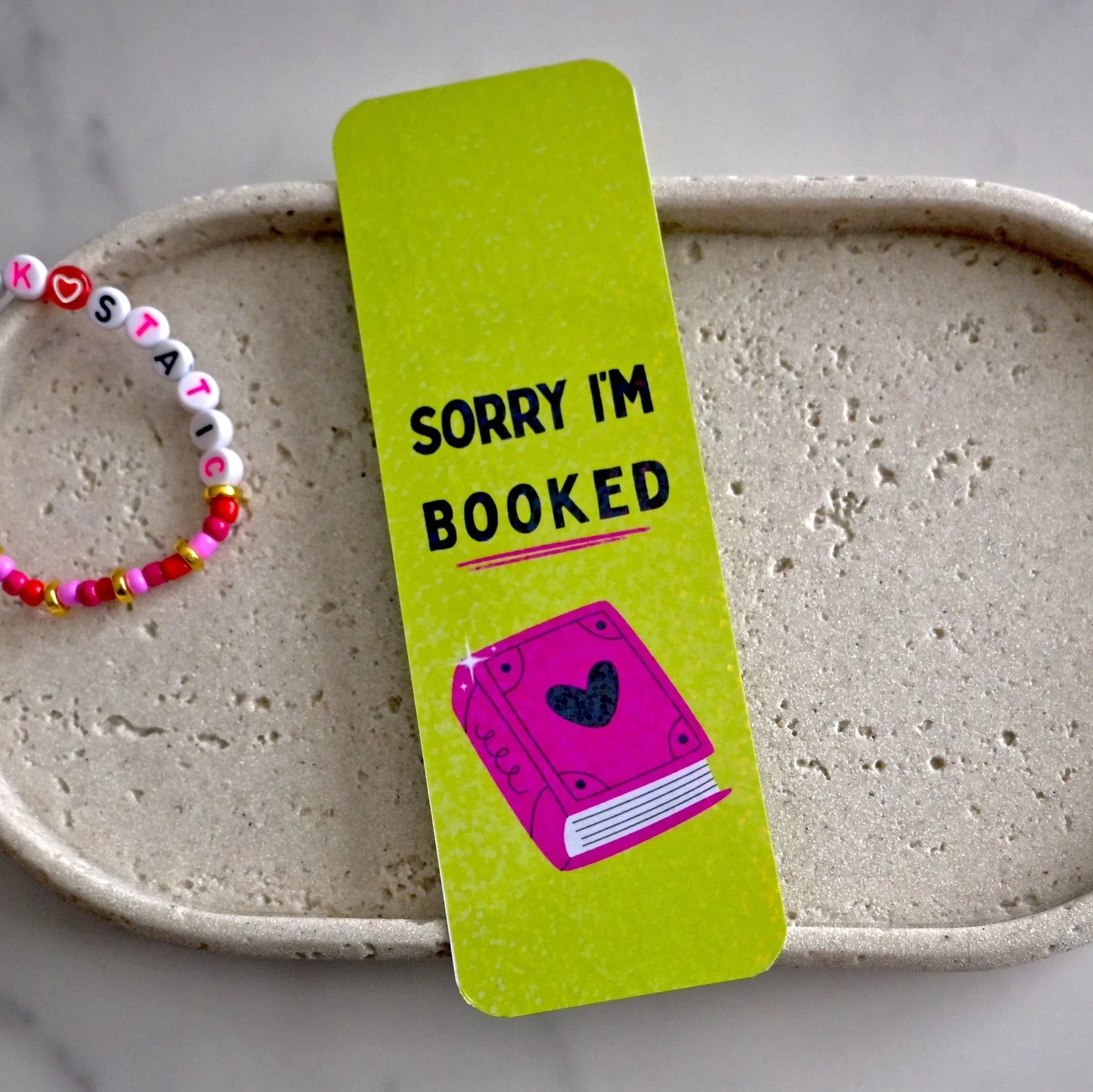 Green 'Sorry I'm Booked' bookmark with a pink book illustration on a ceramic dish with a colorful bracelet.