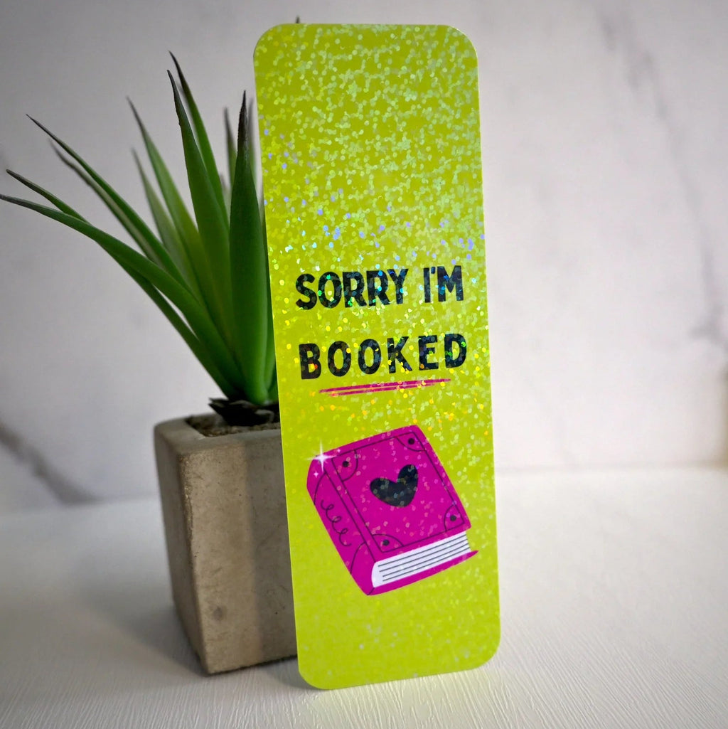 Green bookmark with 'Sorry I'm Booked' text and a pink book illustration, placed next to a plant on a light surface.