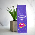 Purple bookmark with 'Talk Bookish To Me' text and lips graphic next to a plant on a light background