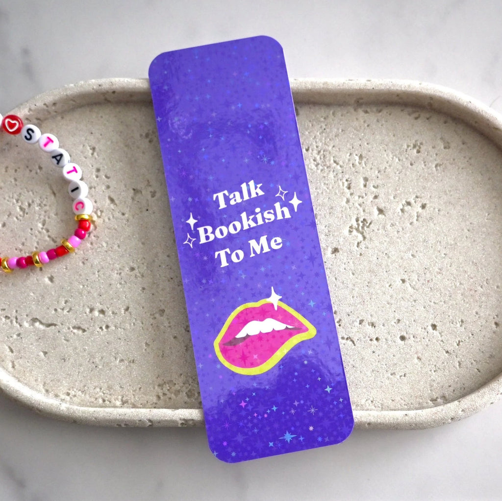 Purple bookmark with 'Talk Bookish To Me' text and lip graphic on a ceramic dish with a bracelet.