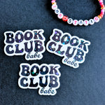 Three 'Book Club Babe' stickers on a dark fabric background with a colorful beaded bracelet.