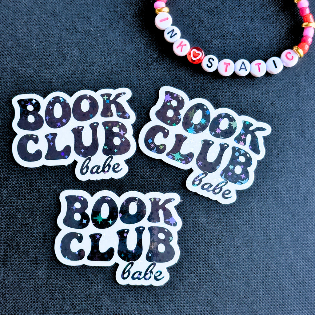 Three 'Book Club Babe' stickers on a dark fabric background with a colorful beaded bracelet.