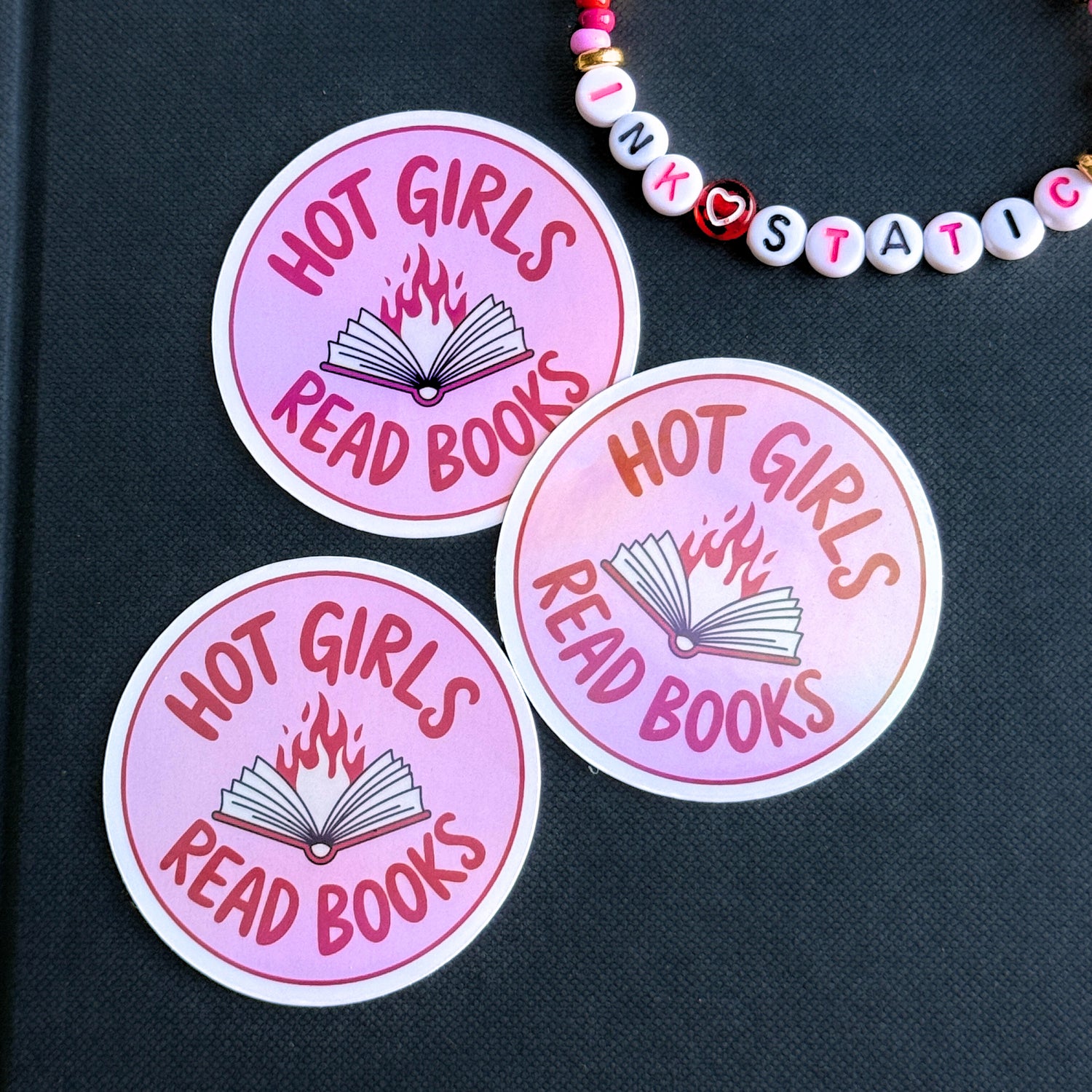 Three pink stickers with 'Hot Girls Read Books' text on a black surface, with a beaded necklace in the background.