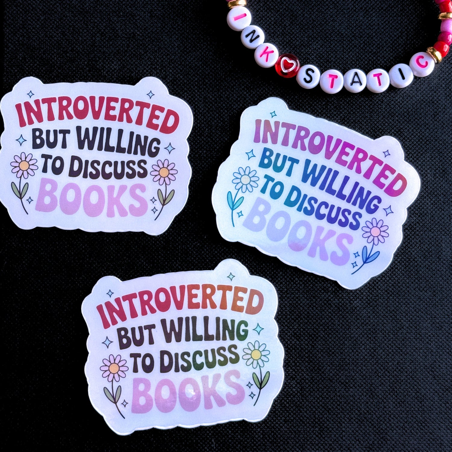Three 'Introverted but willing to discuss books' stickers on a black background with a beaded bracelet.