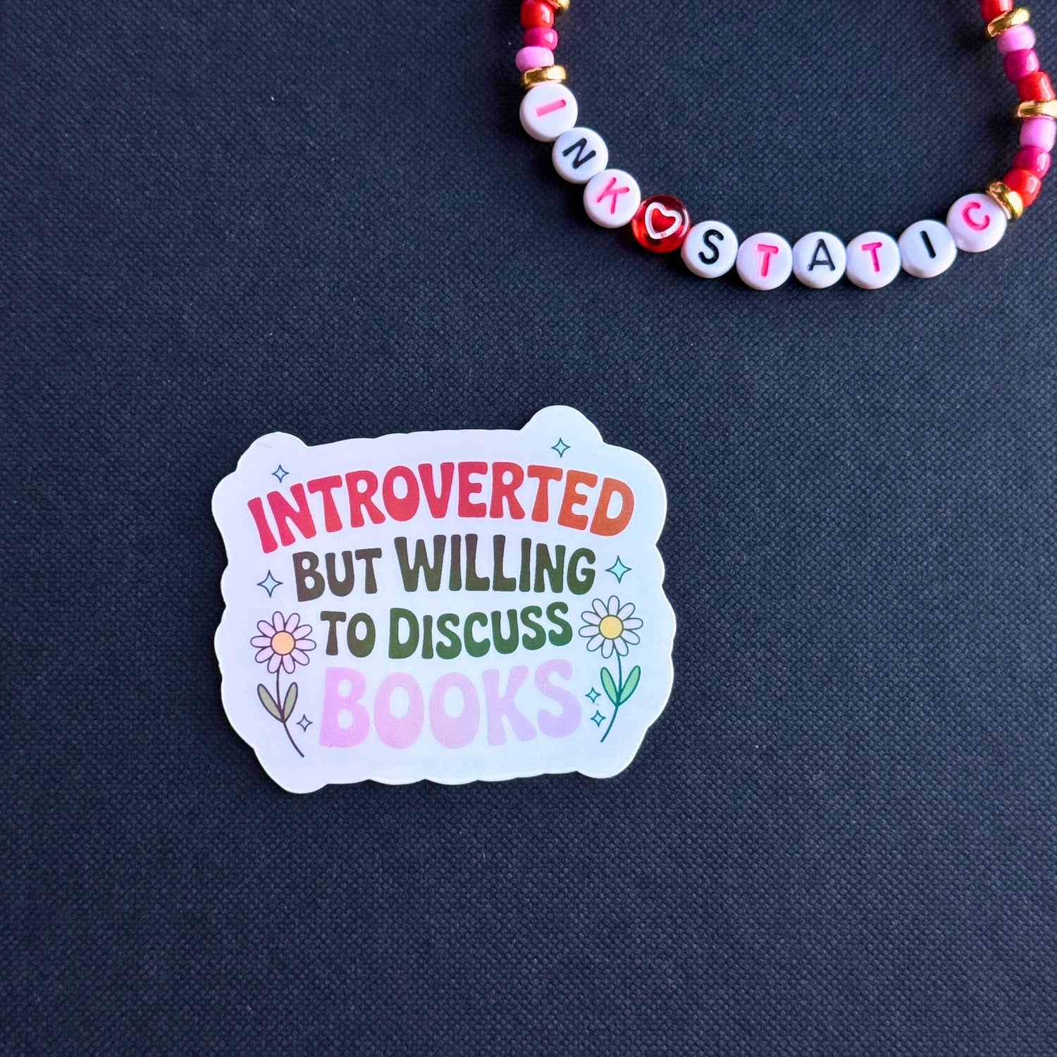 Sticker with 'Introverted but willing to discuss books' text on a dark blue background with a beaded bracelet.