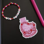 Black notebook with a pink pen, colorful bracelet, and decorative sticker on a gray background