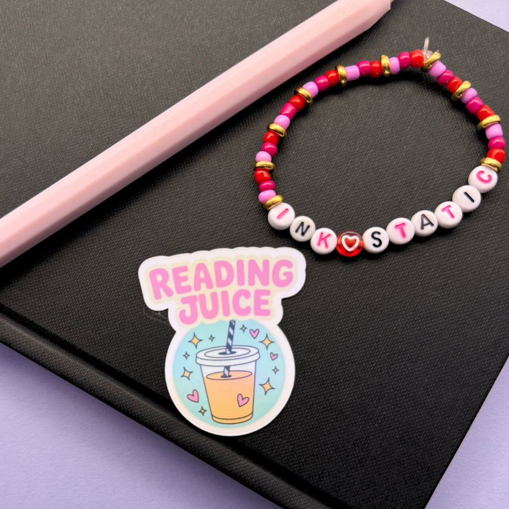 Colorful beaded bracelet and 'Reading Juice' sticker on a black surface with a pink pen.