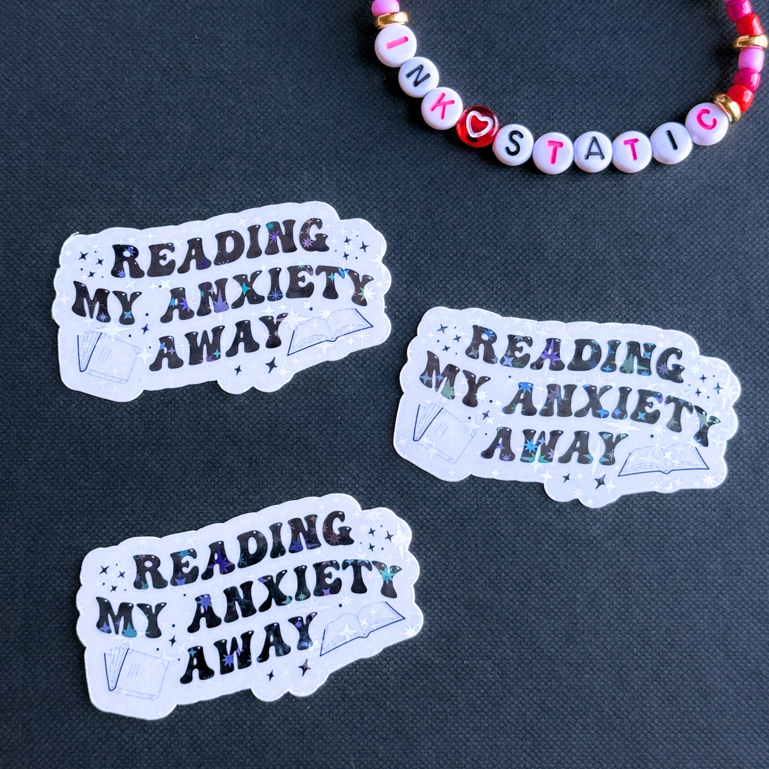 Three 'Reading My Anxiety Away' stickers on a dark fabric background with a beaded bracelet.