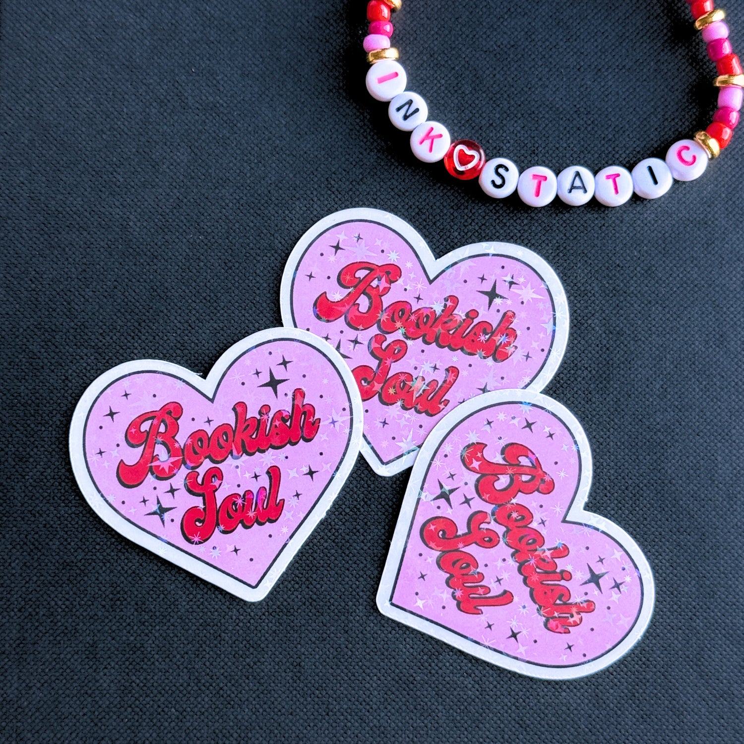 Three heart-shaped 'Bookish Soul' stickers on a dark background with a beaded bracelet.