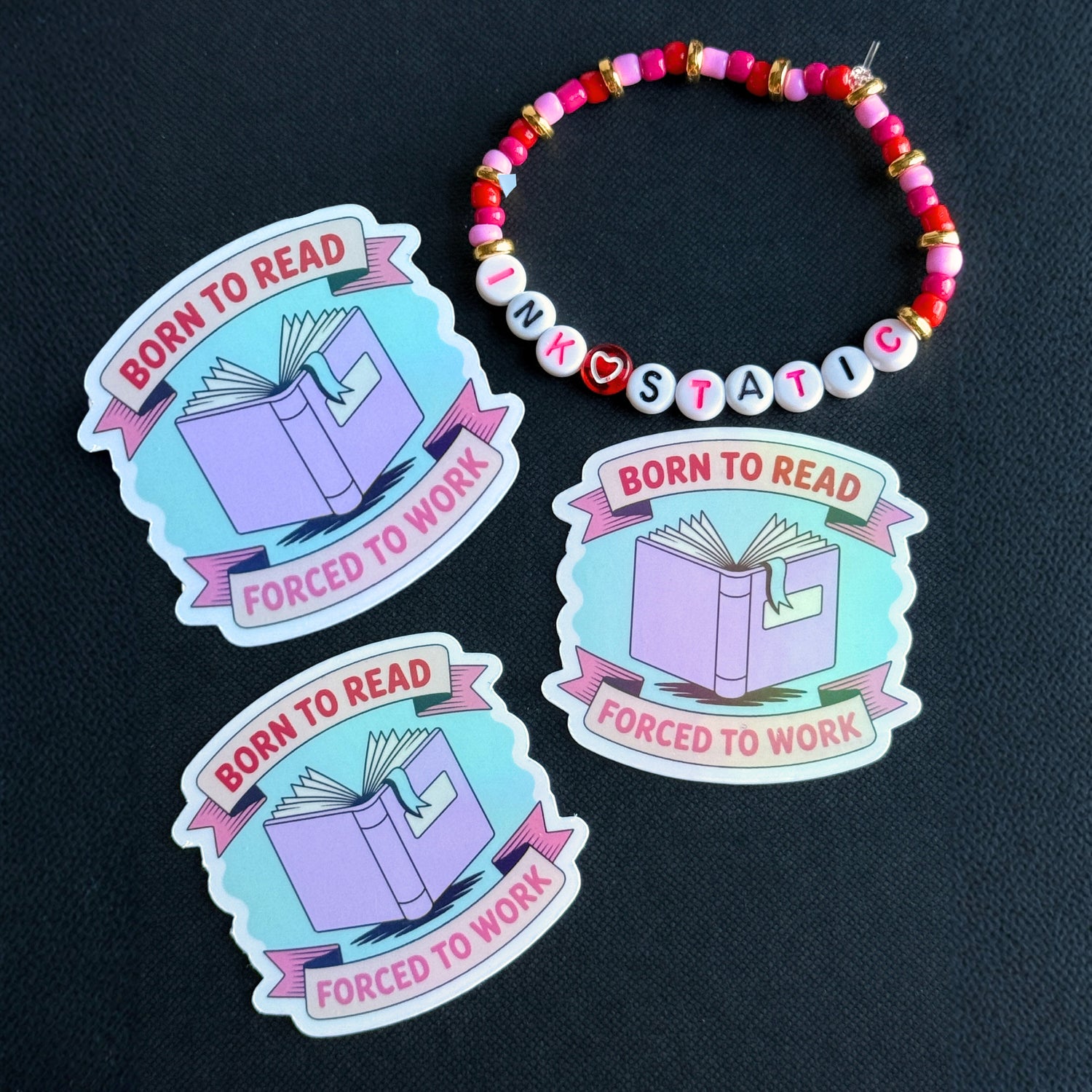 Three 'Born to Read, Forced to Work' stickers with a colorful beaded bracelet on a dark background.